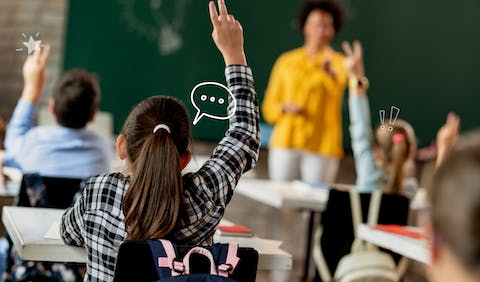 A classroom of students raising their hands with the teacher out of focus in the background