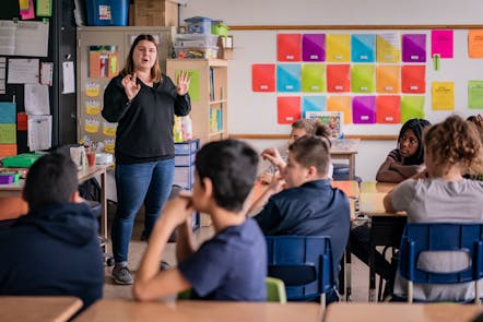 Teacher standing at front of a classroom and explaining something to the students who are sitting at their desks