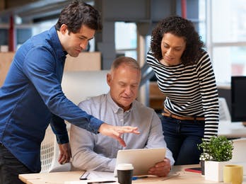 Two teachers standing explaining something on the tablet of a man sitting